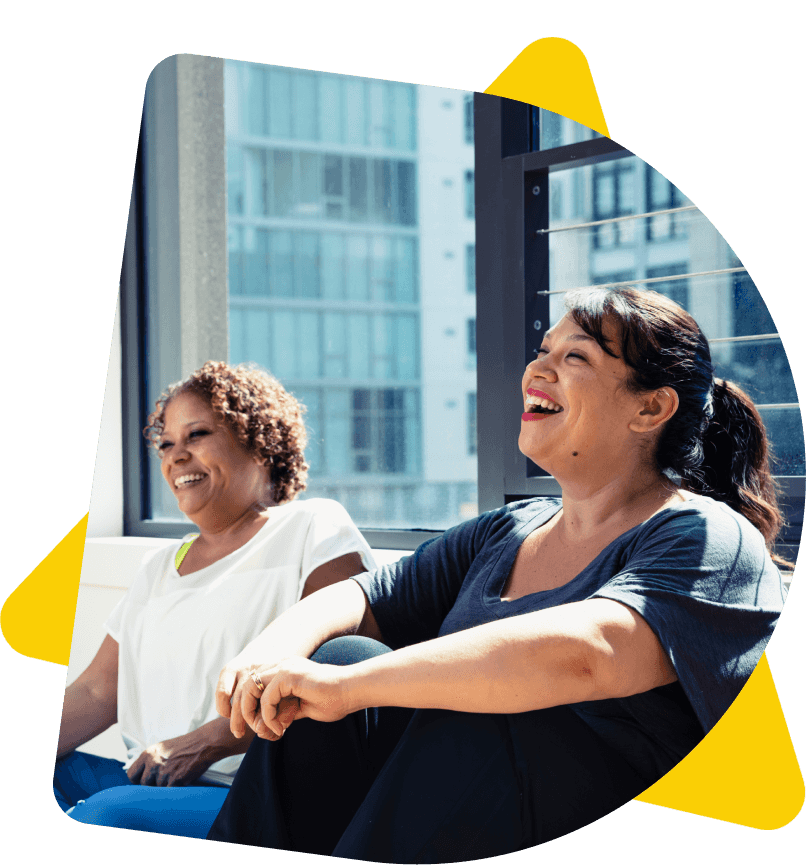 Two happy women laughing together, sat against a window with natural light flooding through