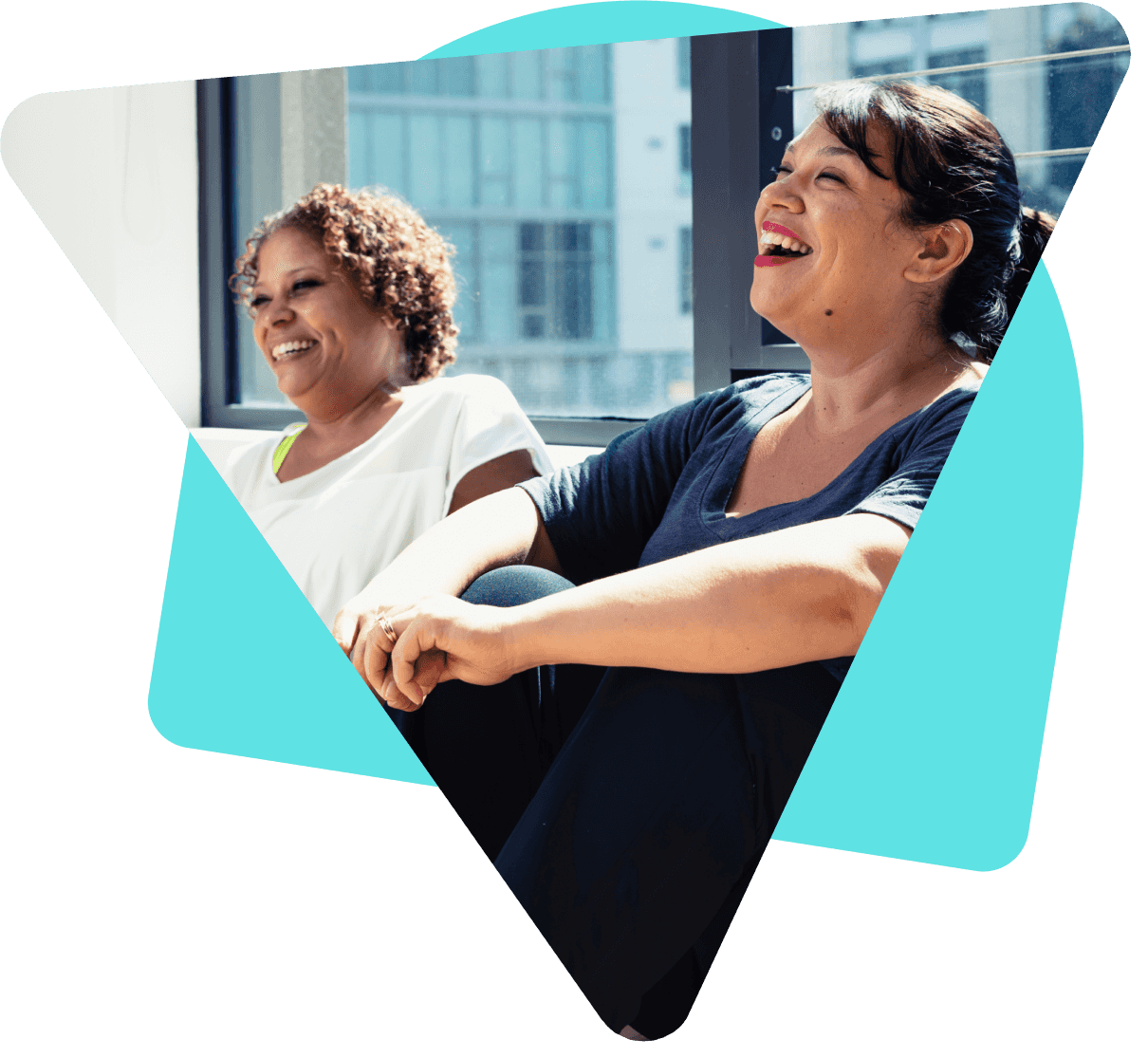 Two happy women laughing together, sat against a window with natural light flooding through