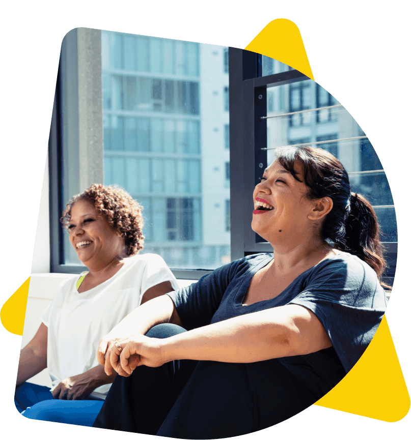 Two happy women laughing together, sat against a window with natural light flooding through